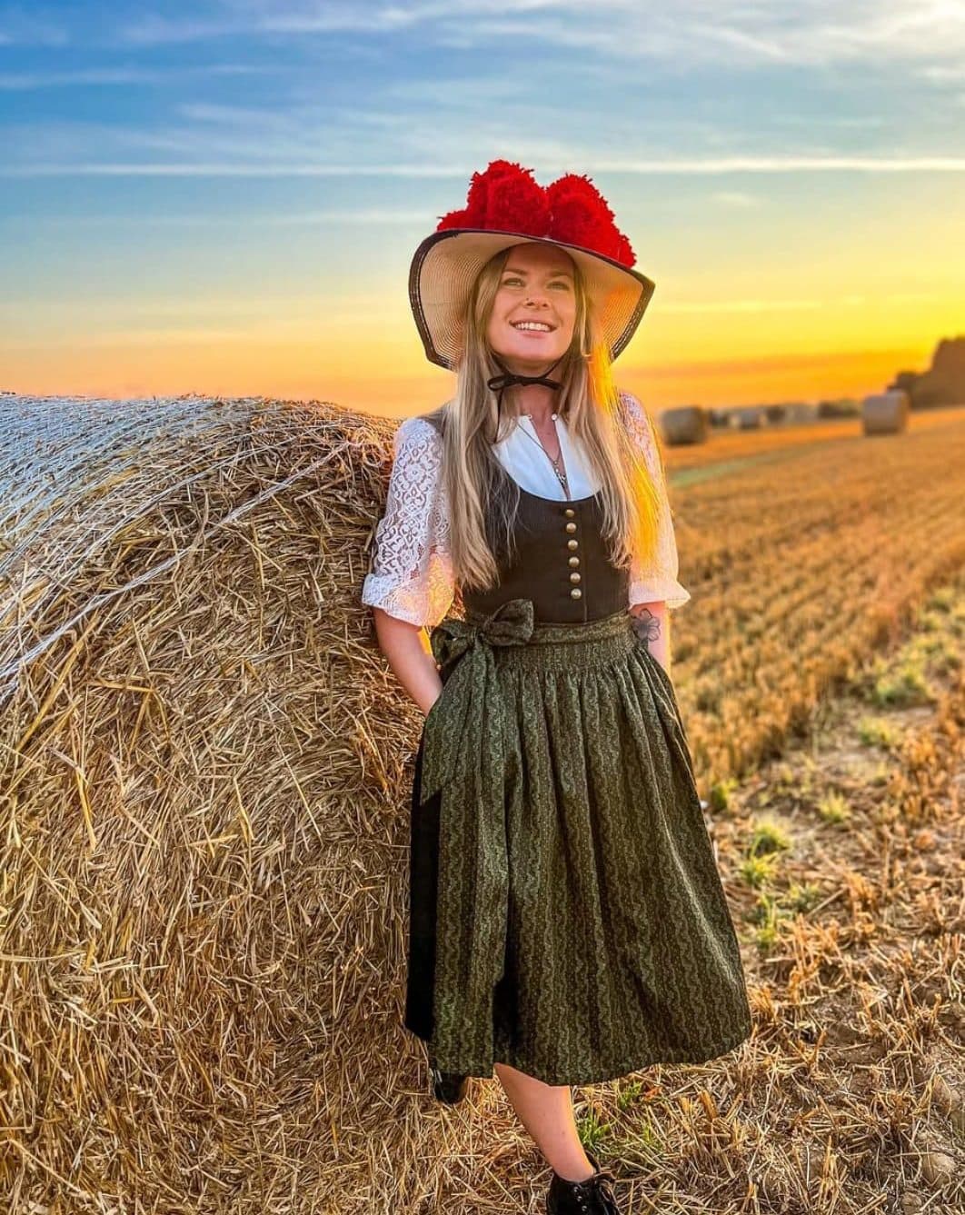 Mary lehnt in Dirndl und Bollenhut an einem Strohballen, hinter ihr geht die Sonne über einem abgeernteten Feld unter.