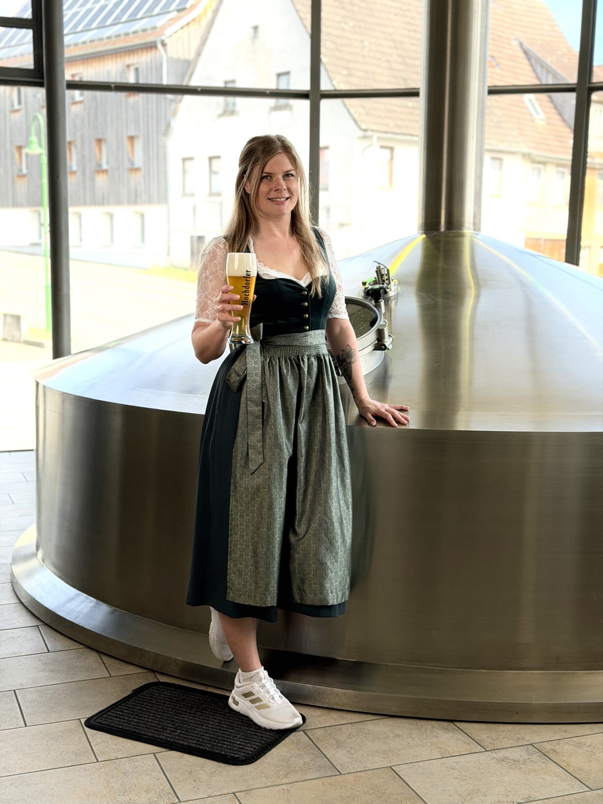 Mary steht im Dirndl mit einem Weizenglas helles Bier neben einem Sudkessel aus Edelstahl, dahinter große Fenster.